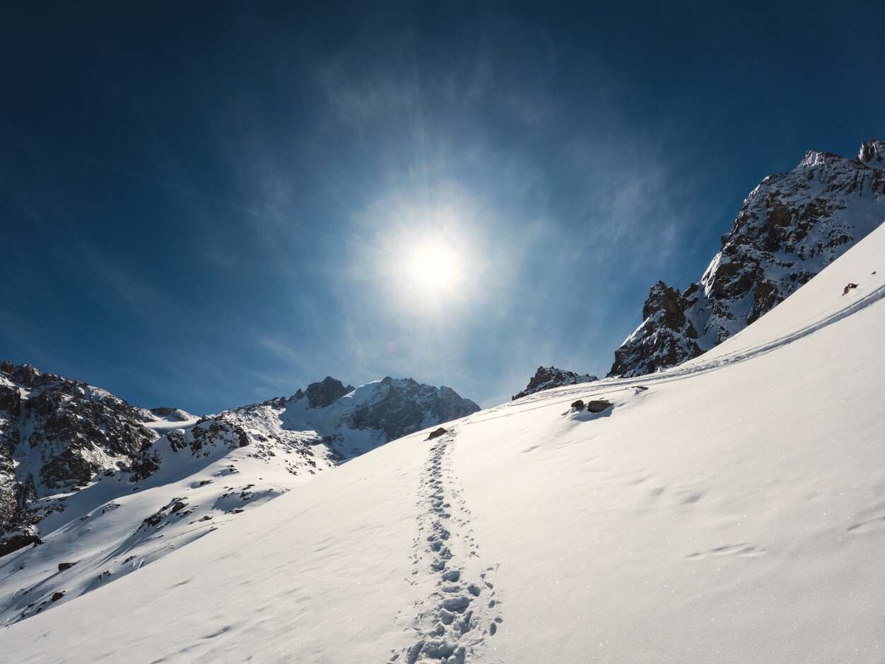 La ladera de una montaña nevada bajo un sol brillante, con huellas que conducen hacia picos rocosos, recuerda a los aventureros que deben utilizar la mejor crema solar para la montaña
