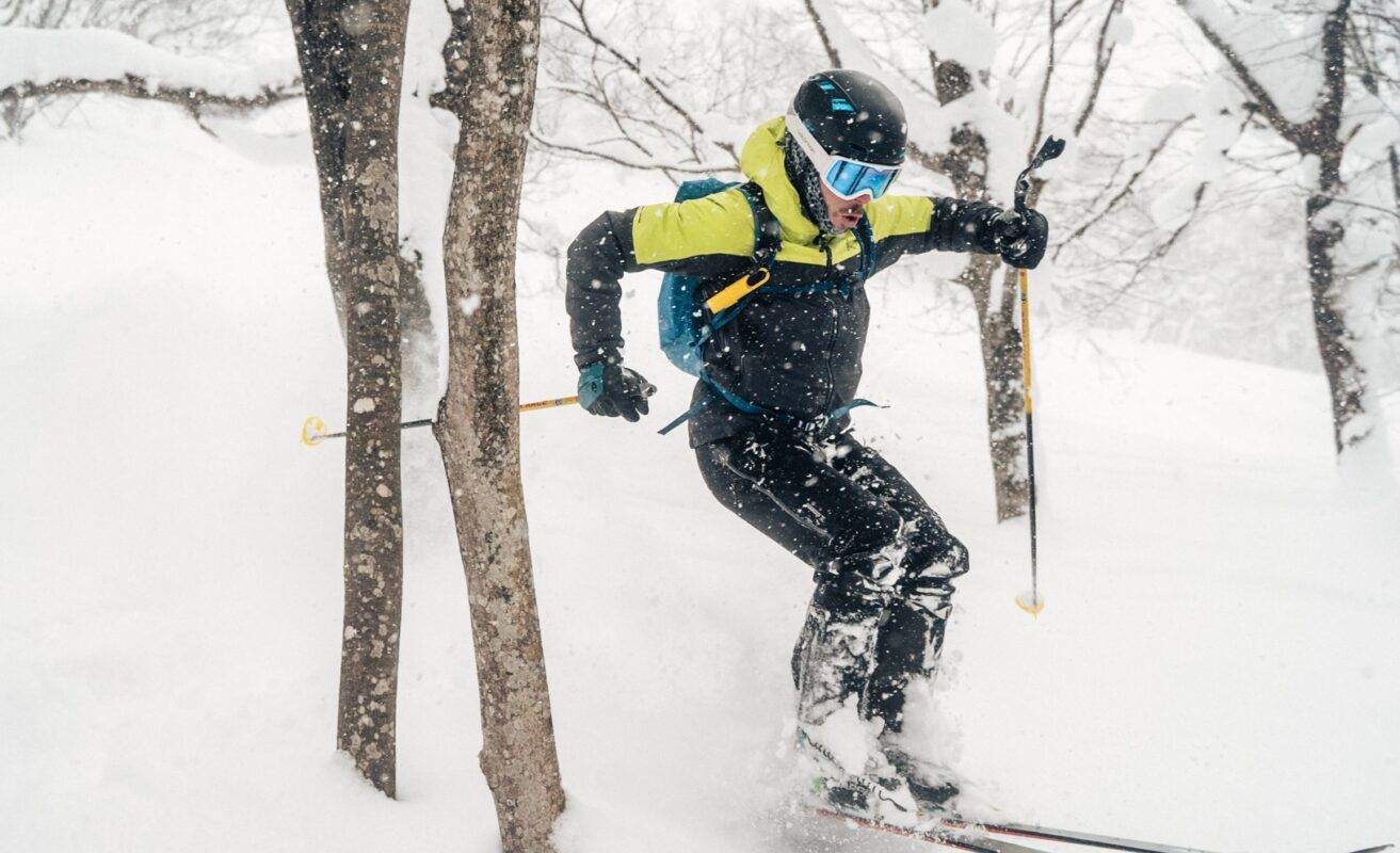 mejor-proteccion-solar-facial Un esquiador con casco negro y chaqueta verde avanza por un bosque nevado, sosteniendo bastones de esquí, mientras la nieve cae a su alrededor.
