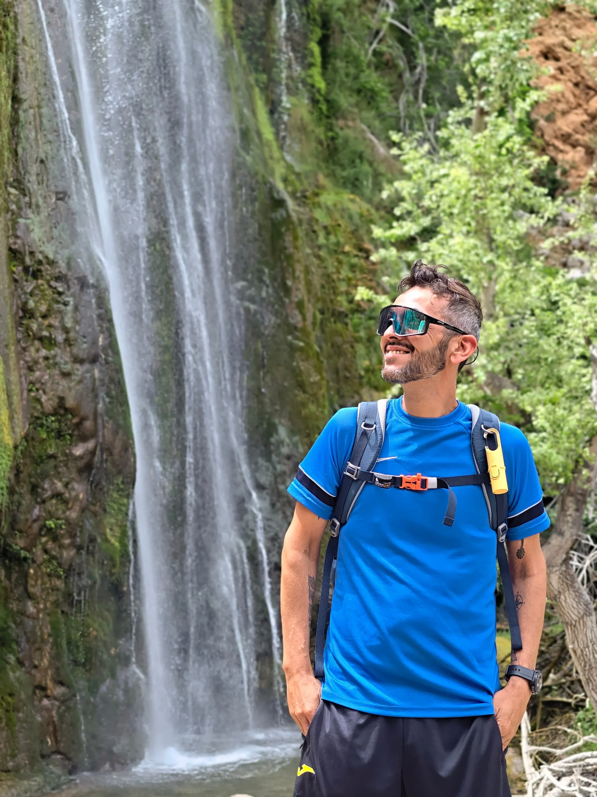 Persona practicando senderismo en montaña con exposición solar, usando mochila y gafas de sol junto a una cascada